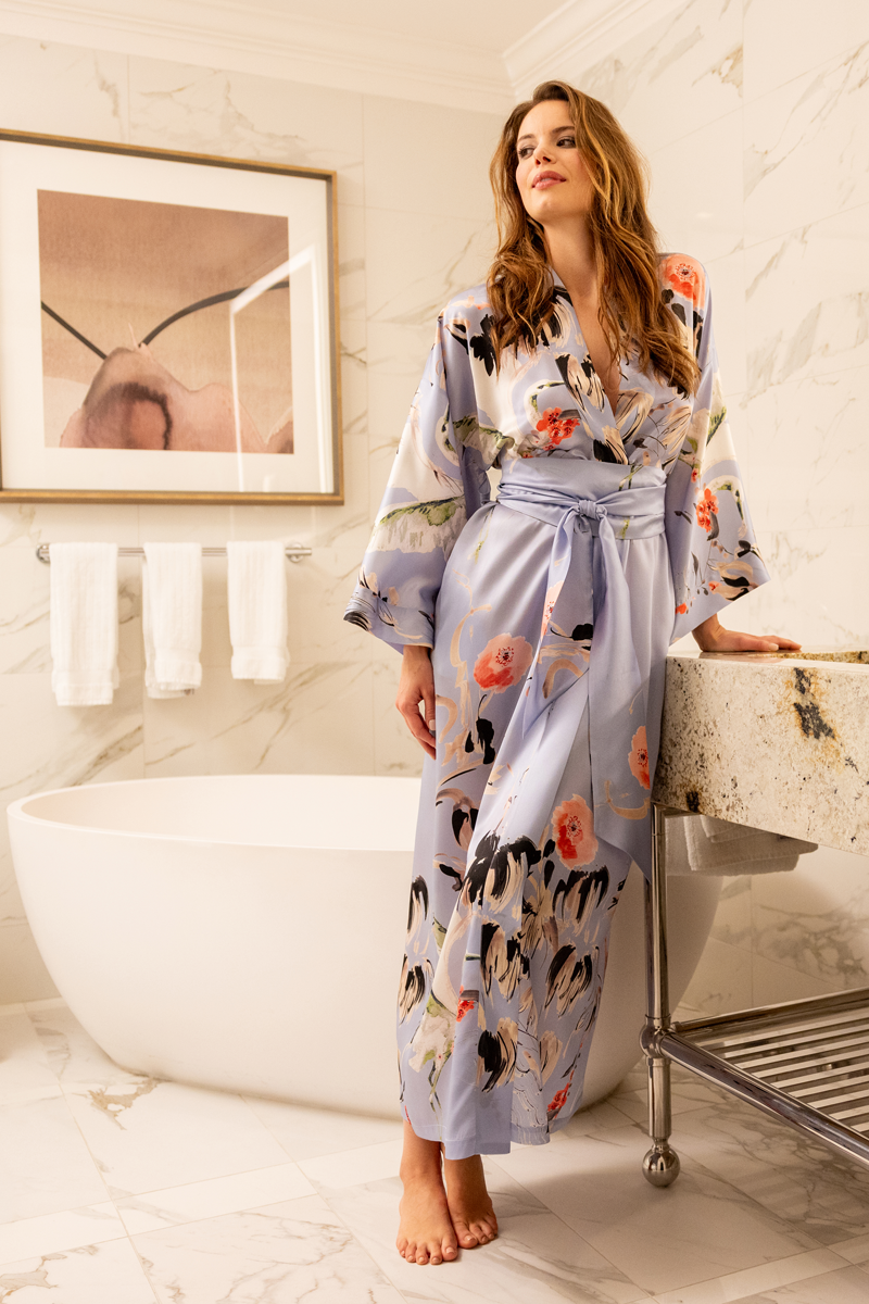 Woman in a floral silk robe standing in a bathroom with a bathtub and framed picture on the wall.