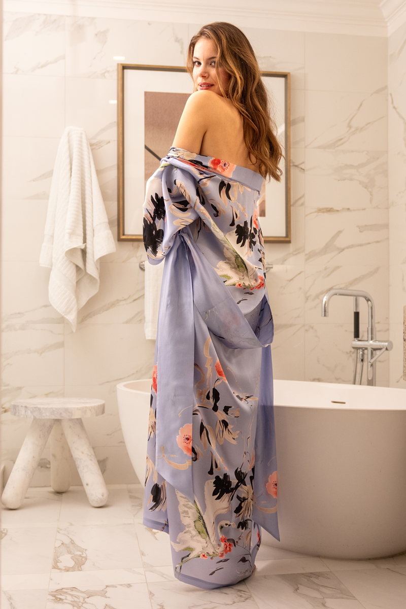 Woman in a floral silk robe standing in a bathroom with a bathtub and framed picture on the wall.